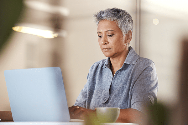 A mature woman with short hair working on a laptop while sitting at a desk with a cup of coffee near them