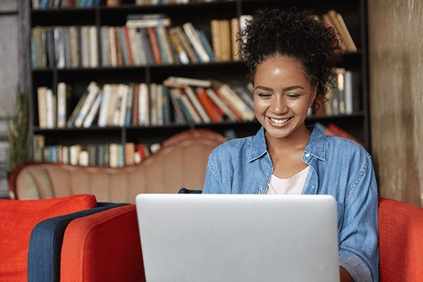 A young adult wearing an opened blue jean button up and white shirt typing on a silver laptop while sitting on a red couch. Behind her is a large bookshelf filled with books.