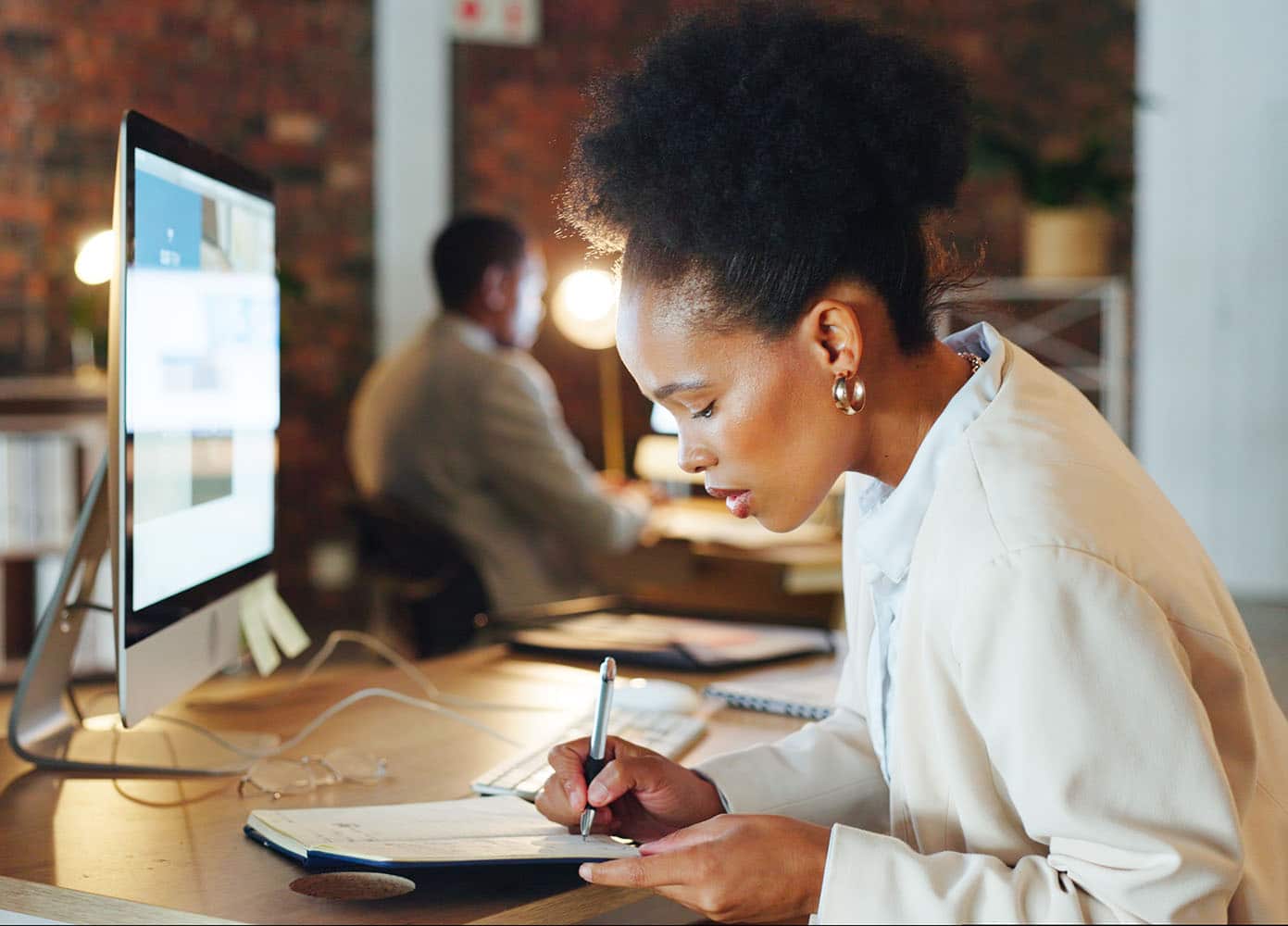 A woman wearing a white long sleeved sweater sitting at a desk writing in a notebook in front of a desktop computer