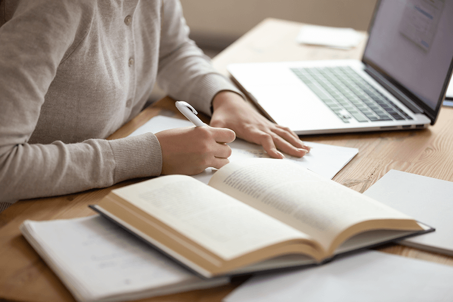 A author working at a desk, writing on paper next to their laptop and an open book