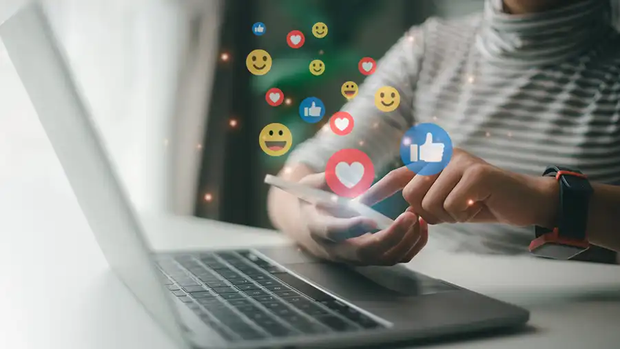 A young person sitting at a desk with their laptop and phone while social media emoticons pop out of the phone screen